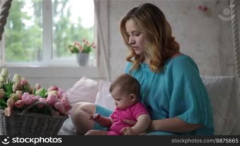 Side view of attractive young mother with her cute infant child sitting on bed in bedroom. Adorable baby girl choosing tulip flowers from basket and playing with flower while family spending leisure at home. Slow motion. Steadicam stabilized shot.
