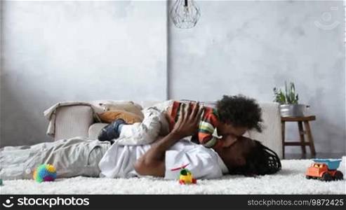 Side view of playful African American father lifting curly mixed race toddler son up while lying on floor at home. Handsome dad with dreadlocks playing with his lovely boy in the living room at home.