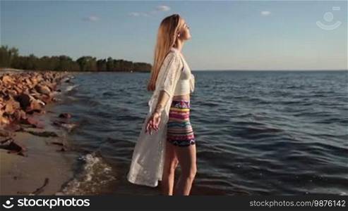 Side view of sensual long blonde hair female enjoying freedom and life in glow of beautiful sunset while standing barefoot on seaside. Blissful girl raising arms feeling free, relaxed and happy on the beach at sunset. Slow motion. Stabilized shot.