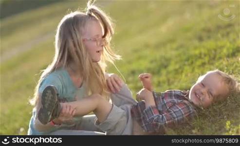 Side view of two playful children having fun on a green field in the park. Lovely blonde sister in eyeglasses kissing the belly of her toddler brother, kids fooling around and laughing. Excited siblings enjoying time together outdoors.