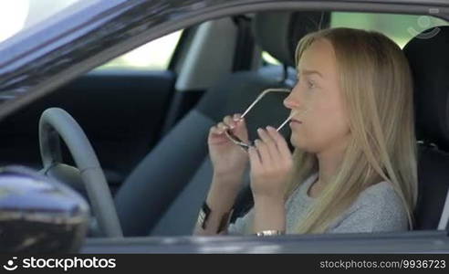 Side view of young female sitting in driver's seat, putting sunglasses on and fastening seat belt.