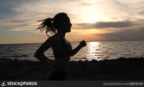 Side view of young fit female with ponytail running and exercising in the evening on the seaside. Silhouette of sporty runner woman jogging on the beach in twilight time. Slow motion. Steadicam stabilized shot.