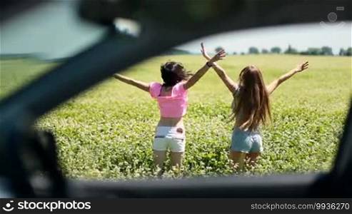 Side view through the open car window of cute young happy women jumping with their hands up in flowers field, while traveling by car in countryside