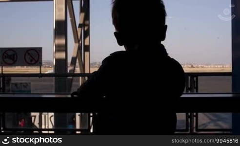 Silhouette of a young boy looking through a window in the airport to the passengers passing by and airplanes