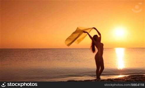 Silhouette of a young woman in a bikini holding a cloth over her head posing at sunset
