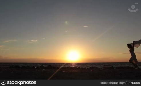 Silhouette of happy woman running with flying scarf on the beach against dramatic sky during sunset. Carefree long hair female in summer dress with flying scarf in hands jogging on the sea shore in glow of orange setting sun. Slow motion.