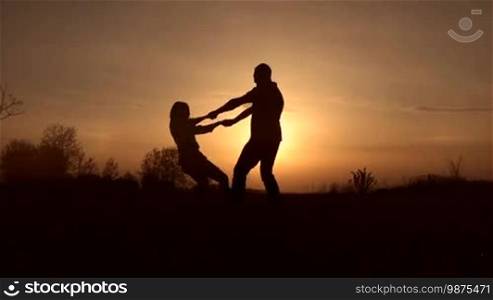 Silhouettes of a couple in love holding hands and spinning around in the glow of an awesome sunset. The joyful couple shares the best moments of togetherness, having fun while spending a great time outdoors at sunset. Slow motion. Steadicam stabilized shot.