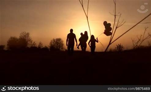 Silhouettes of happy family with children walking into the rays of setting sun in meadow. Rear view. Cheerful young mother carrying toddler son, daughter holding balloons and father watching beautiful sunset. Slow motion. Steadicam stabilized shot.