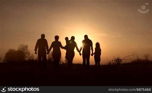 Silhouettes of three generation family holding hands walking in the meadow in the glow of a beautiful sunset. Rear view. Happy family with two kids enjoying an amazing view of the setting sun outdoors in spring. Slow motion. Steadicam stabilized shot.
