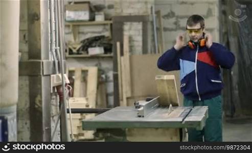 Skilled carpenter cutting piece of wood in his woodwork workshop, using circular saw and wearing safety goggles and earmuffs with other machinery in the background. Craftsman cutting wooden plank with circular saw in workshop.