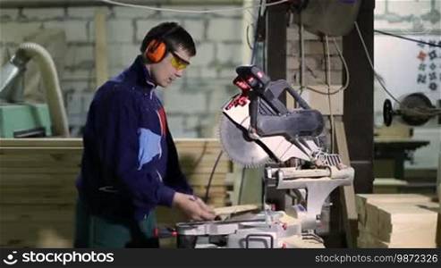 Skilled carpenter in protective workwear measuring wooden plank with ruler and pencil for cutting. Master joiner sawing board with circular saw machine in woodworking shop.