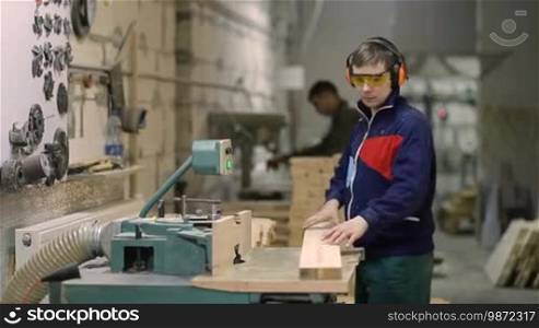Skilled craftsman cutting wooden plank with circular saw in workshop. Carpenter wearing protective glasses and earmuffs working with circular saw machine and decorating wooden board at the workplace.