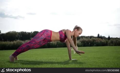 Slim pretty woman with ponytail planking on the green grass on a sunny day. Full length of a sporty young female doing the side rotating plank yoga pose in fitness on grass outdoors. Side view