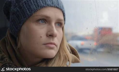 Slow motion and close-up shot of a young woman with a frustrated and sad look. She is traveling by bus in the rainy city and staring through the window