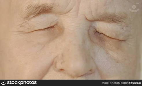 Slow motion and close-up shot of elderly woman's face. She is opening her eyes, looking at the camera, and closing them again