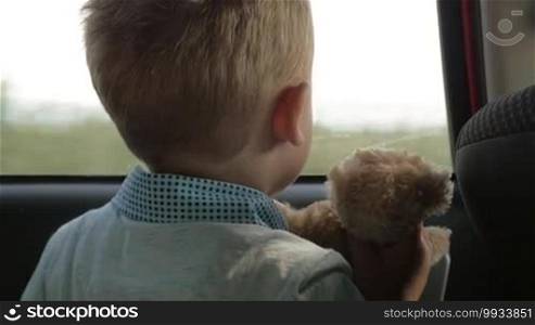 Slow motion close-up shot of a little boy traveling by car with favorite toy bear. He is hugging his soft friend and they are enjoying the view through the window
