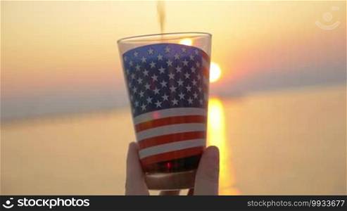 Slow motion close-up shot of pouring soda into the glass with American flag and woman having refreshing drink on the background of sea and sunset