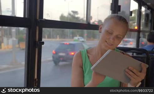 Slow motion of a young blonde woman working with a tablet computer during a bus ride in the city. She is sitting by the window with a view of road traffic