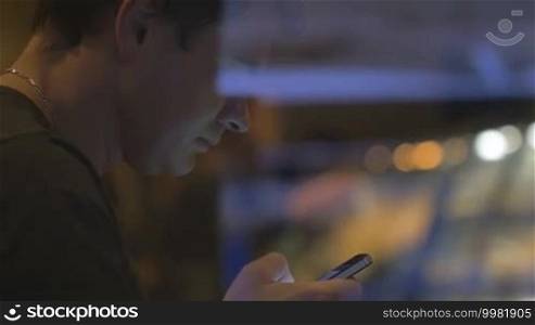 Slow motion of a young man using a smartphone to surf the internet or browse social media while sitting in a cafe in the evening. View through the glass