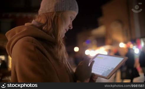 Slow motion of a young woman with a touchpad in the evening city. She is typing and sending a message in a chat. Blurred street with lights and people in the background