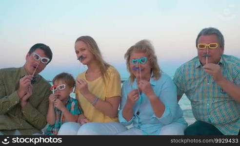 Slow motion of big hipster style family sitting on the beach by the sea. Happy parents, child, and grandparents holding paper glasses and mustache