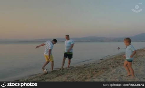 Slow motion of father, son, and grandfather playing football on the beach in the evening. Family team in blue and white striped t-shirts