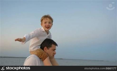 Slow motion of happy little boy sitting on father's shoulders and imitating plane while they walk by the sea