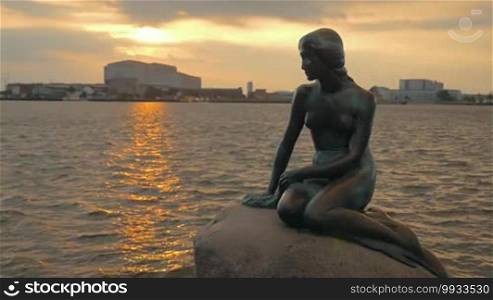 Slow motion of rippling water with golden sun reflection and The Little Mermaid statue on the stone in foreground. Copenhagen, Denmark