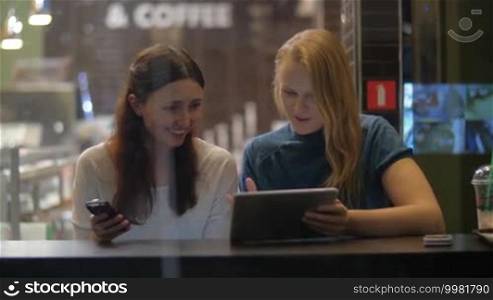 Slow motion of two female friends in a cafe. They are watching something humorous on a digital tablet and laughing. One of the women is also using a cell phone. View through the glass