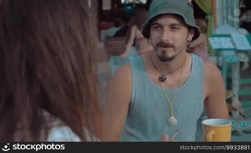 Slow motion of two friends sitting in outdoor cafe. Young man with beard wearing bucket hat listening to his smoking long-haired old chap