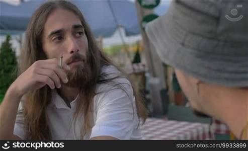 Slow motion of two male friends sitting in an outdoor cafe. They are smoking and having a conversation. One man has long hair and a beard, while the other is wearing a bucket hat.