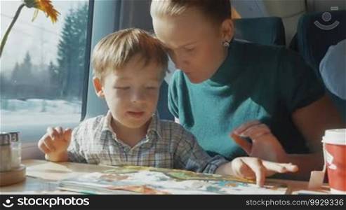 Slow motion of young mother and little son looking at pictures in children's book while traveling by train