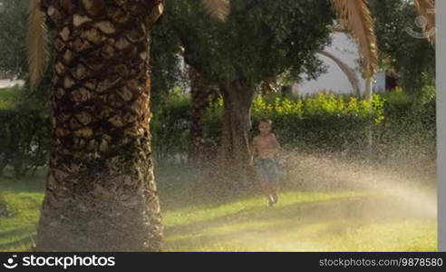 Slow motion shot of a boy having fun running on the lawn with automatic sprinkler watering the grass in the garden