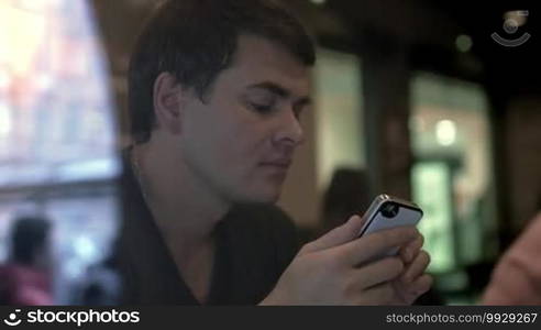 Slow motion shot of a man sitting in a cafe and writing messages on a smartphone. Shot is taken through the cafe's window.