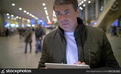 Slow motion shot of a man watching something on a tablet PC, he's standing in an airport or railway terminal.