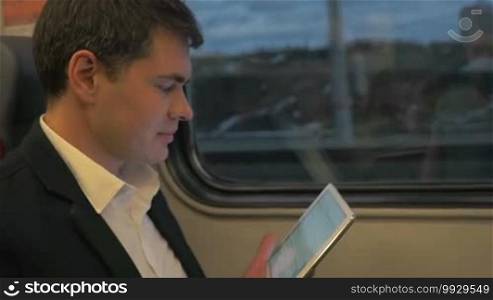 Slow motion shot of a man wearing a business suit traveling by train. He is reading something funny on a tablet and laughing.