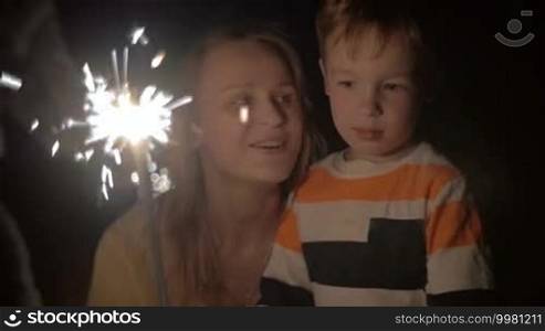 Slow motion shot of a young woman and her little brother looking at sparkles of Bengale fire. Woman is squatting near the boy and tell him something.