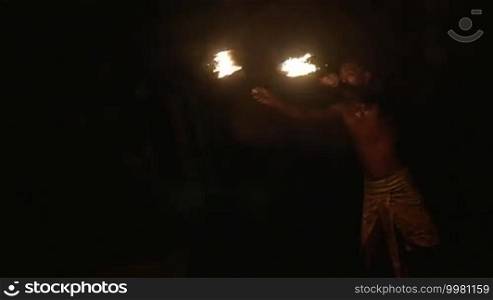 Slow motion shot of an episode of fire show. Dark-skinned artist makes a huge fire explosion with two lighting torches.