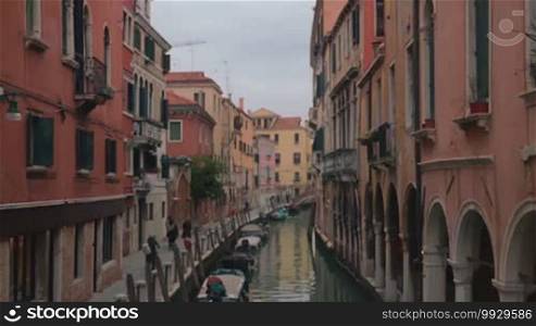 Slow motion shot of common street of Venice: water canal and narrow sidewalk are surrounded with ancient high-rise buildings.