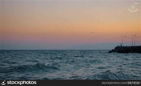 Slow motion shot of waving sea, rocky pier, and lonely seagull flying in the evening sky