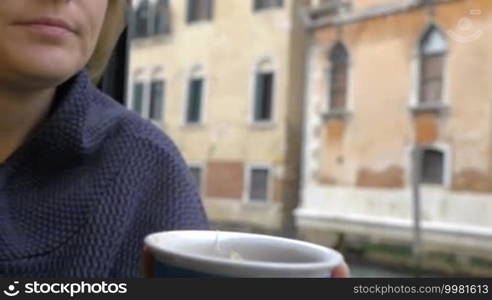 Slow motion, steadicam, and close-up shot of a woman drinking hot tea from a colorful cup sitting by the window at home