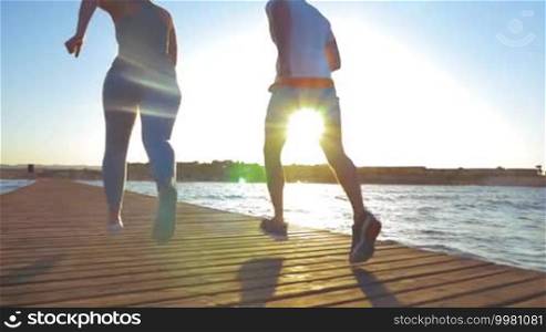 Slow motion steadicam and low angle shot of man and woman running on wooden pier on coastal resort at sunset