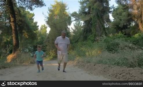 Slow motion steadicam shot of a little boy and grandfather having a run up the road in the forest. Active and healthy outdoor leisure