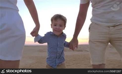 Slow motion steadicam shot of a playful little boy taking away parents' hands and running away. Happy mother and father following him. Family fun on vacation