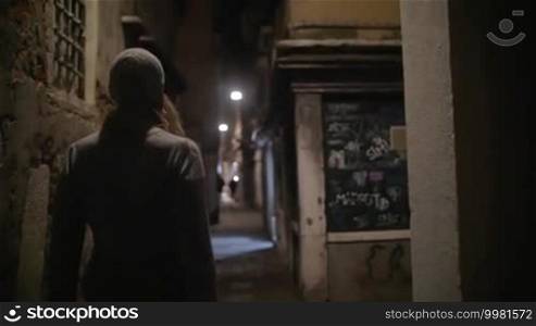 Slow motion steadicam shot of a woman walking along the alleyway with dim light. Night city with worn grungy buildings