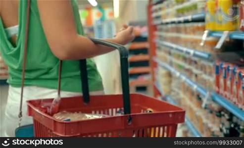 Slow motion steadicam shot of a woman with a plastic shopping cart walking along the shelves in the supermarket. Shopping for food