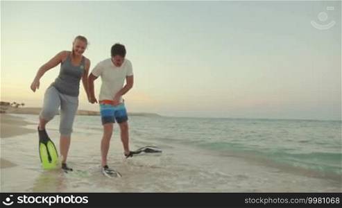 Slow motion steadicam shot of a young man and woman walking in flippers along the seashore. They look funny and clumsy