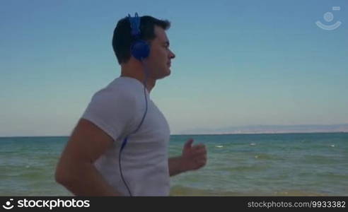 Slow motion steadicam shot of a young man having regular jog training along the coast, sea, and clear blue sky in the background. He is listening to music in headphones and using a smartphone while running