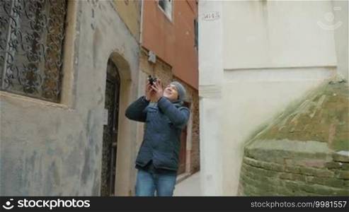 Slow motion steadicam shot of a young man walking among aged worn buildings in Venice and shooting exterior with retro camera