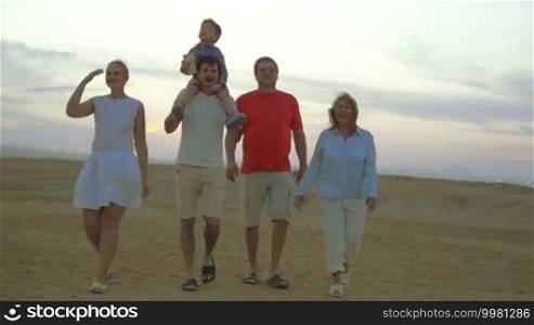 Slow motion steadicam shot of happy big family walking outdoor. Grandparents, young parents and little son riding on father's neck enjoying evening on the beach